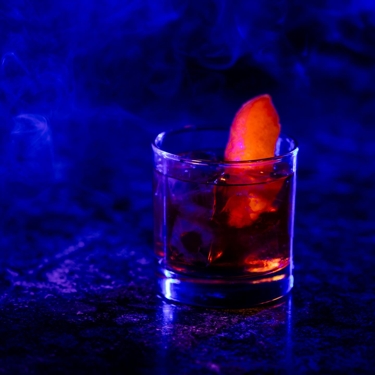 Dark drink with ice, orange slice, and blue smoky background.