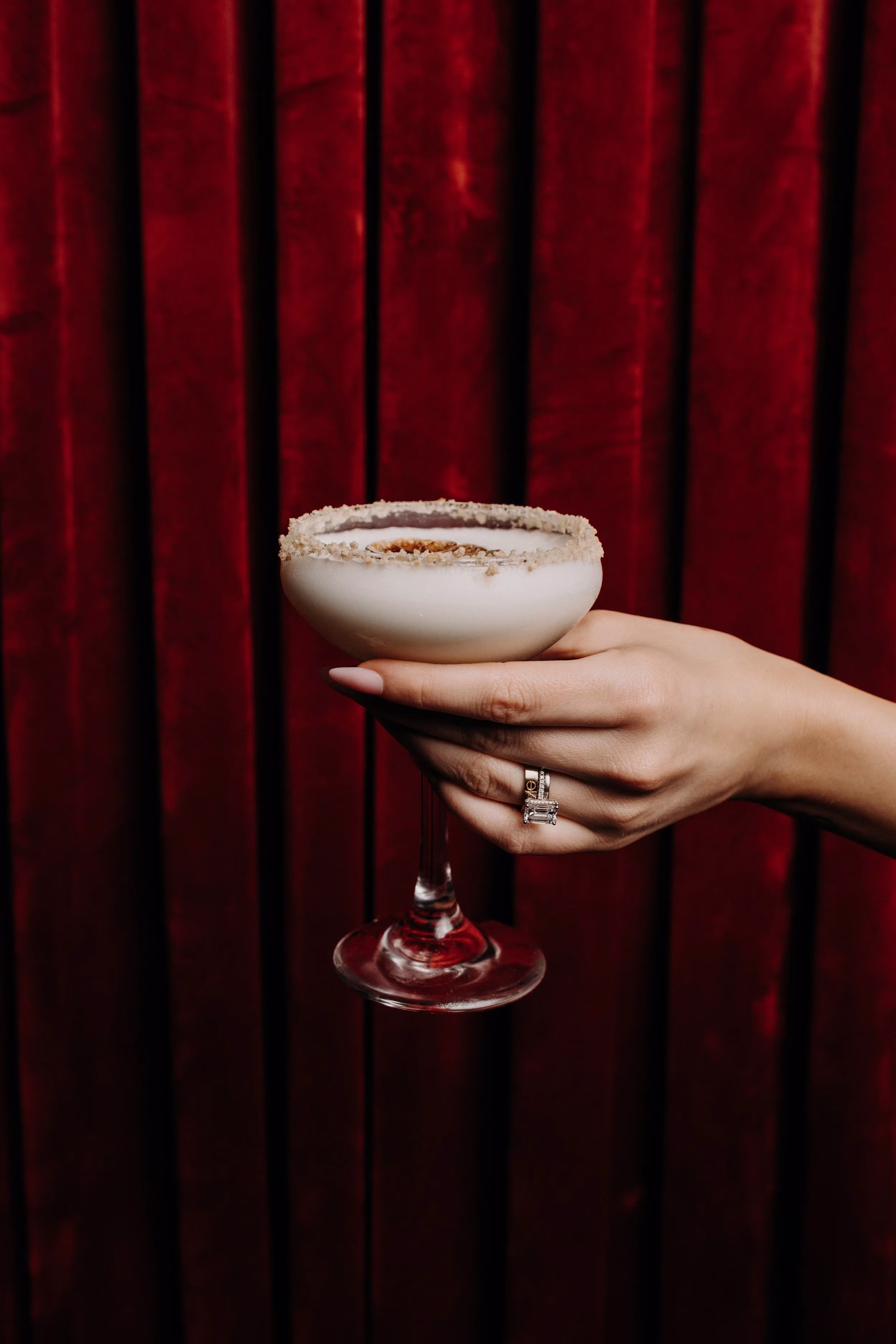 A hand holding a white, sugar-rimmed cocktail against a dark red curtain.
