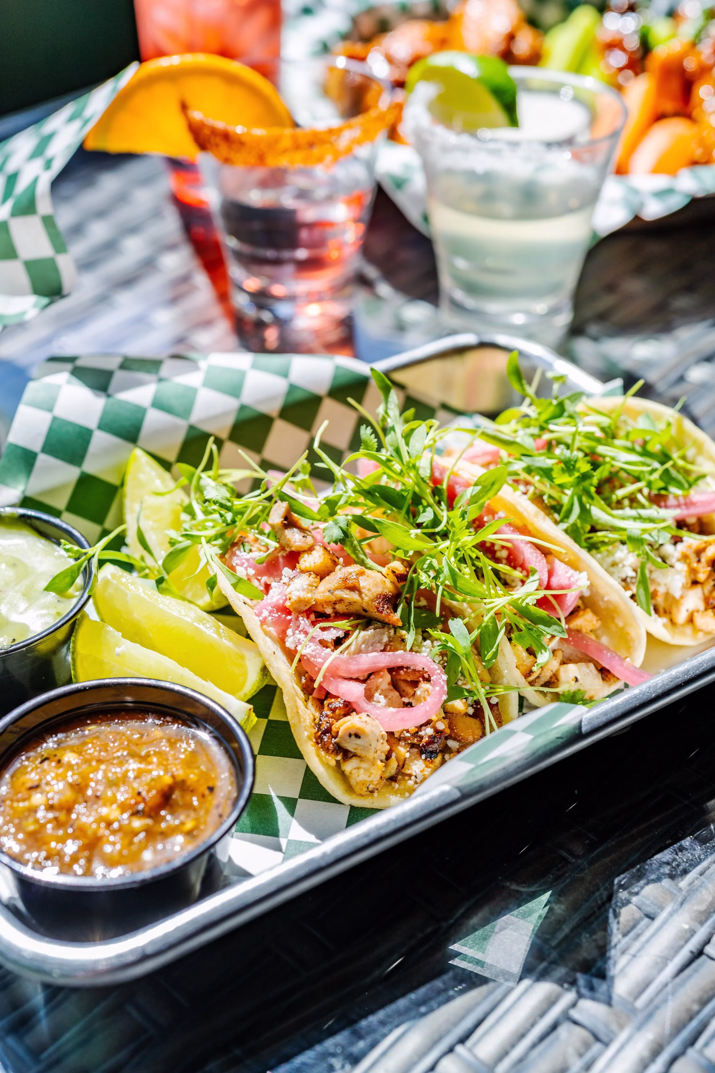Chicken tacos with pickled onions, microgreens, sauces, lime on checkered tray.