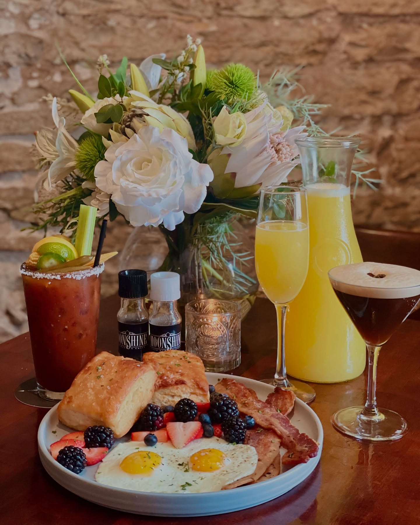 Full brunch meal with eggs, bacon, biscuits, drinks, and flowers.