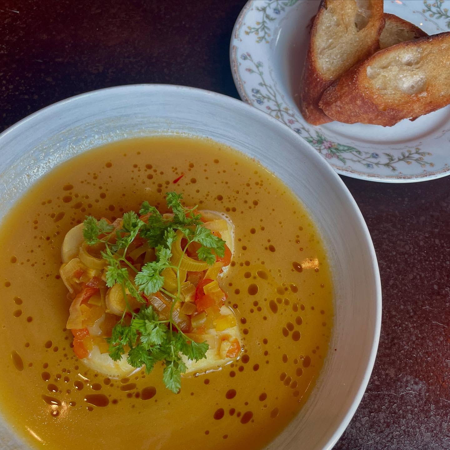 White bowl of yellow broth with garnished main dish, toasted baguette.