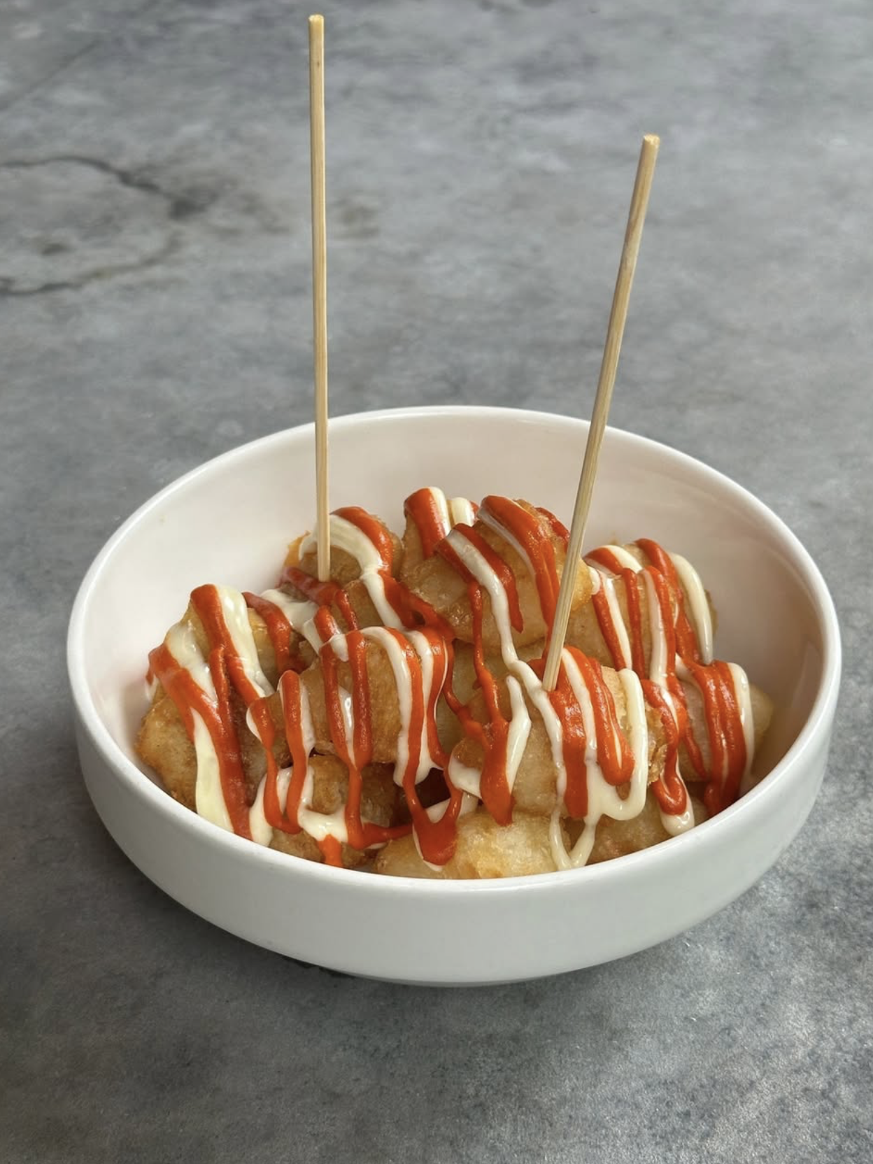White bowl of fried pieces with red and white sauce and skewers.