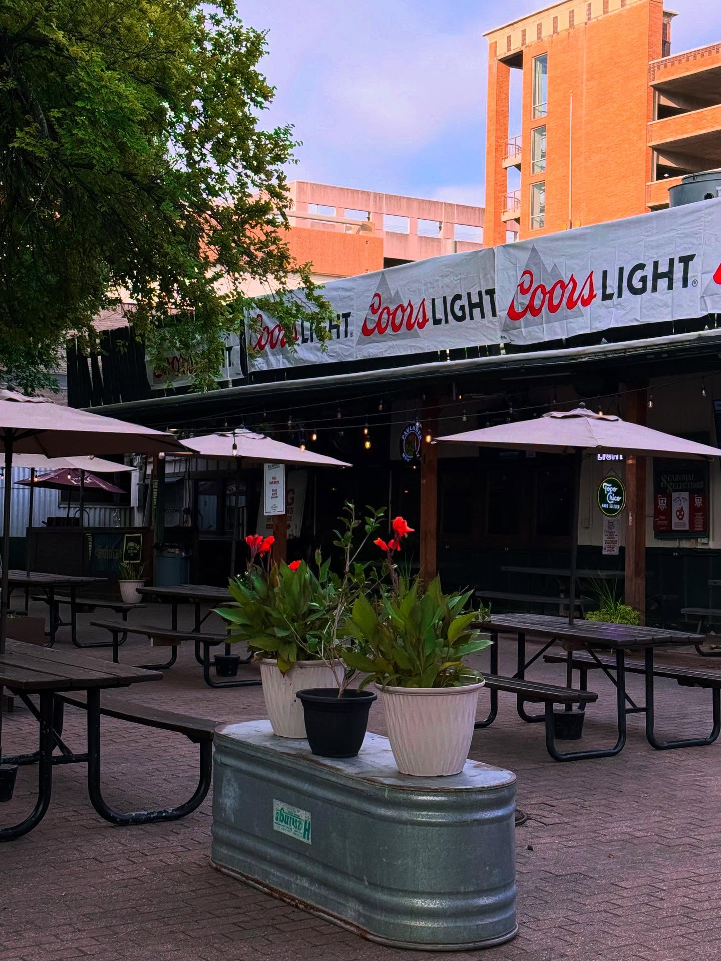 Outdoor patio with picnic tables, umbrellas, potted plants, and Coors Light banner.