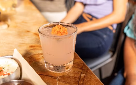 Light pink cocktail garnished with orange flower on a wooden table.