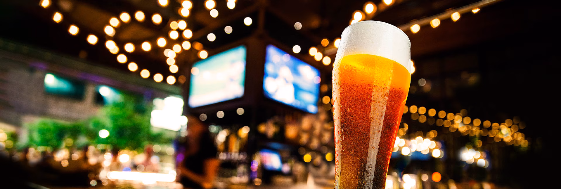 Glass of frothy beer with condensation, blurred string lights and screens.