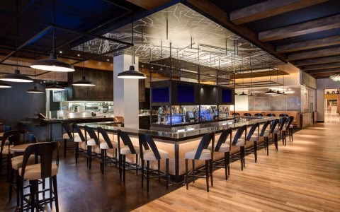Empty modern bar with numerous stools, open kitchen, and wooden floor.