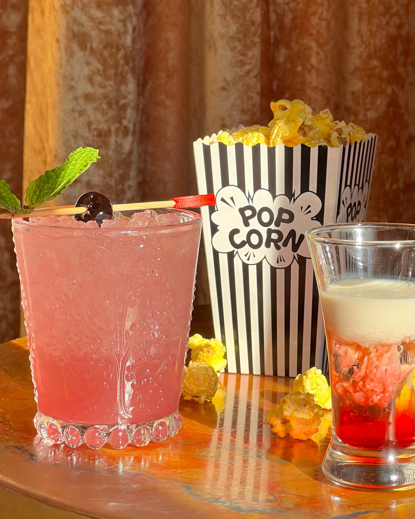 Pink cocktail, popcorn box, and layered shot glass on reflective surface.