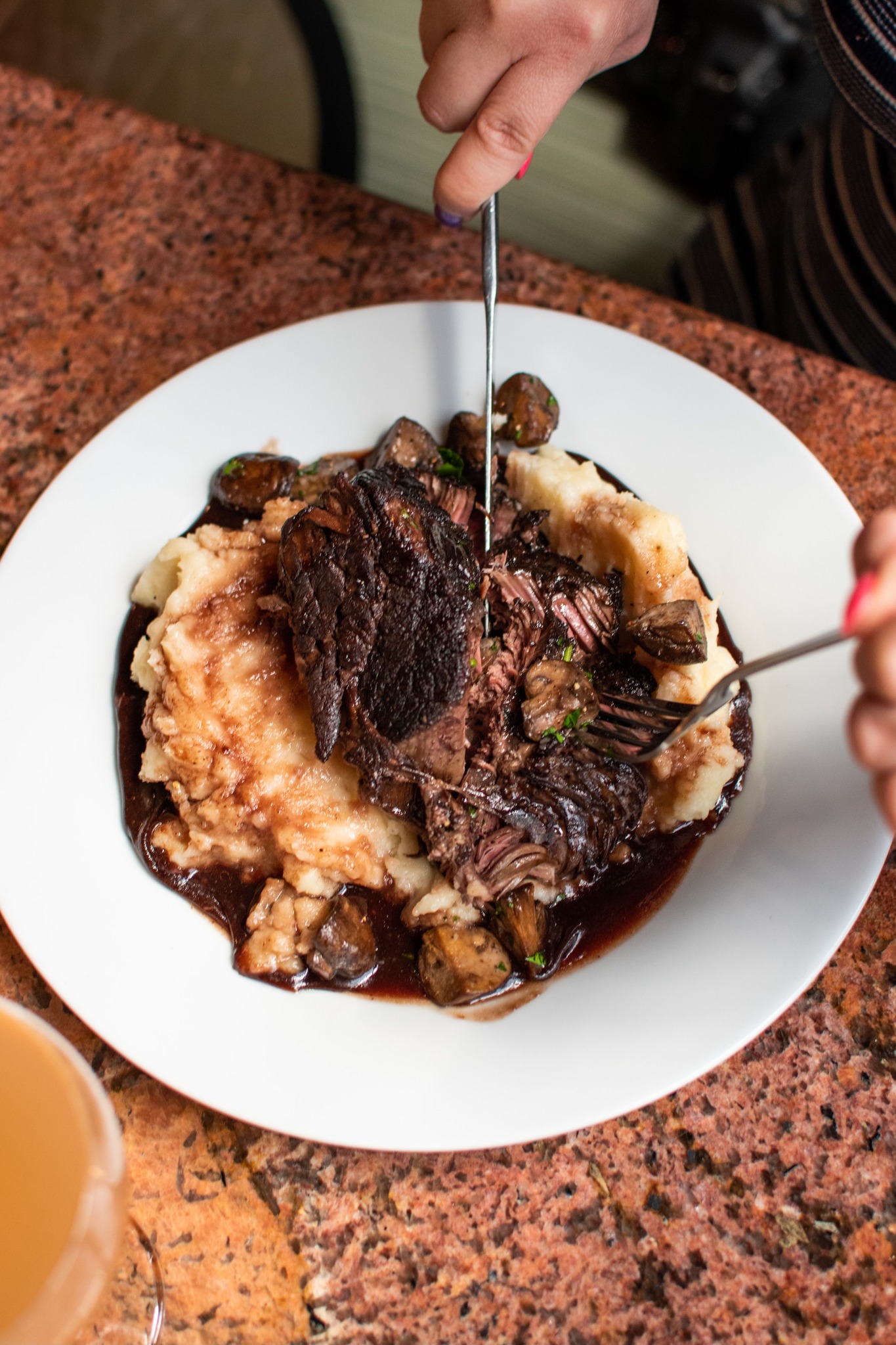 Hands cutting braised beef with mashed potatoes, mushrooms, and gravy.