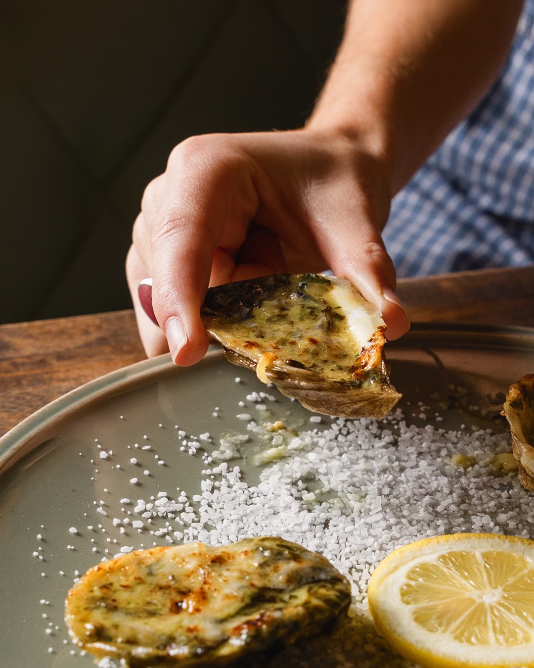 Hand holds a broiled oyster above a plate with salt and lemon.