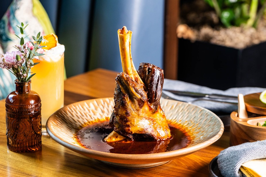 Meat shank in sauce, with a drink and flowers on a restaurant table.
