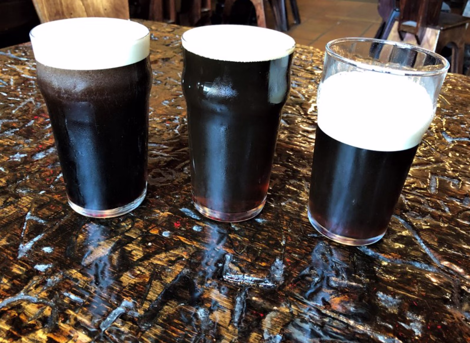 Three dark beers with white foam on a textured wooden table.