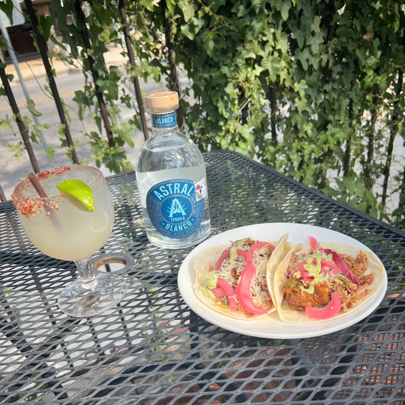 Margarita, Astral Tequila Blanco bottle, and two tacos on outdoor table.