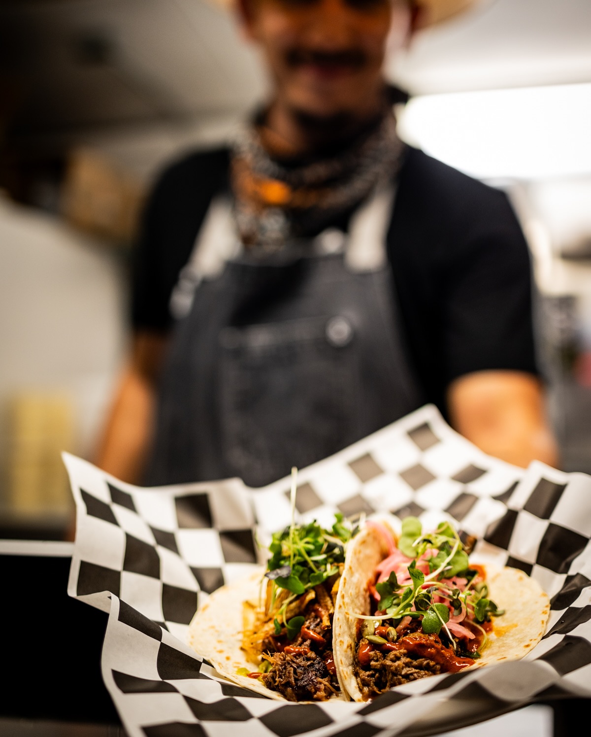 Two tacos with meat, microgreens, and toppings, presented by a person.