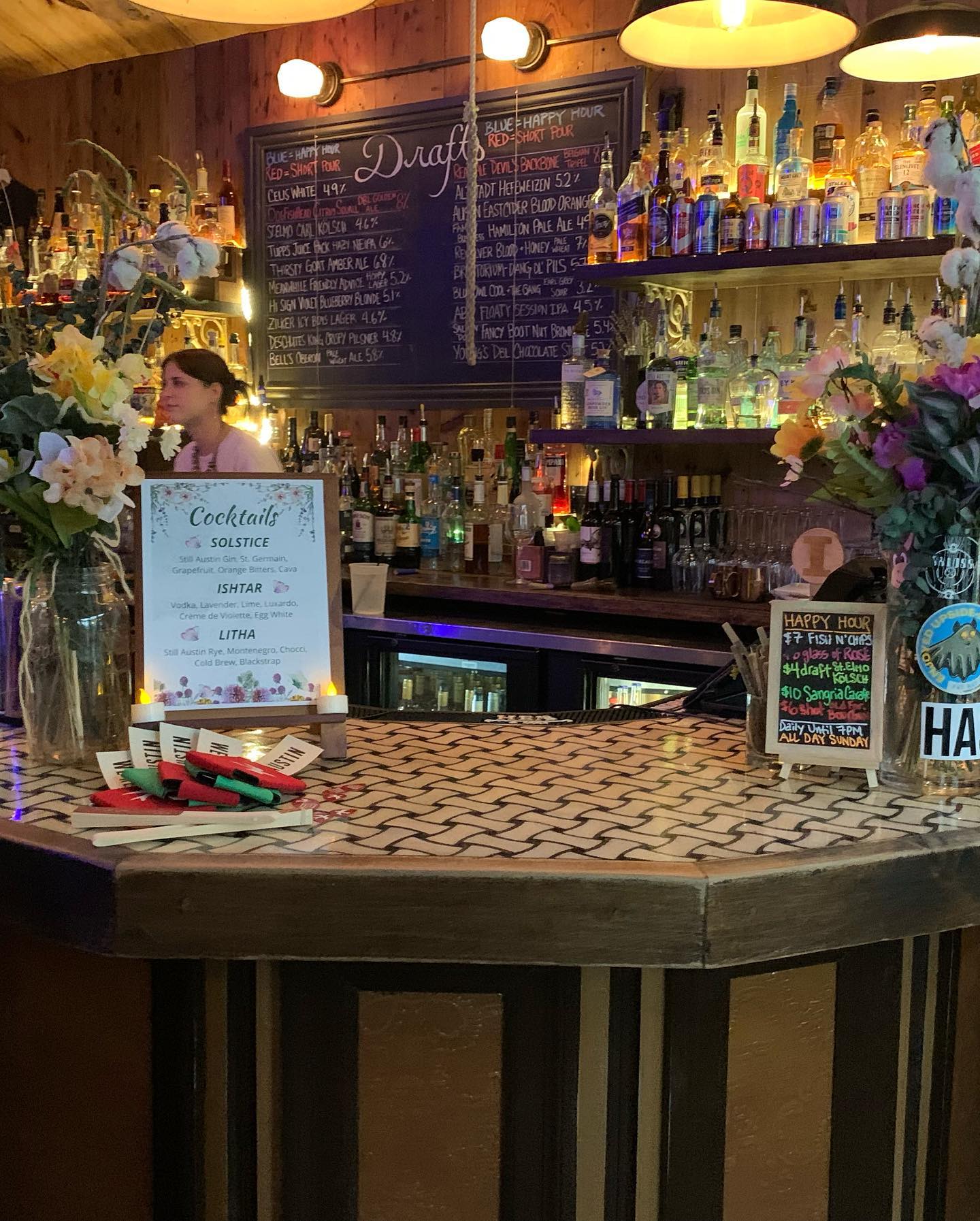 Bartender behind a well-stocked bar with menus and flowers.