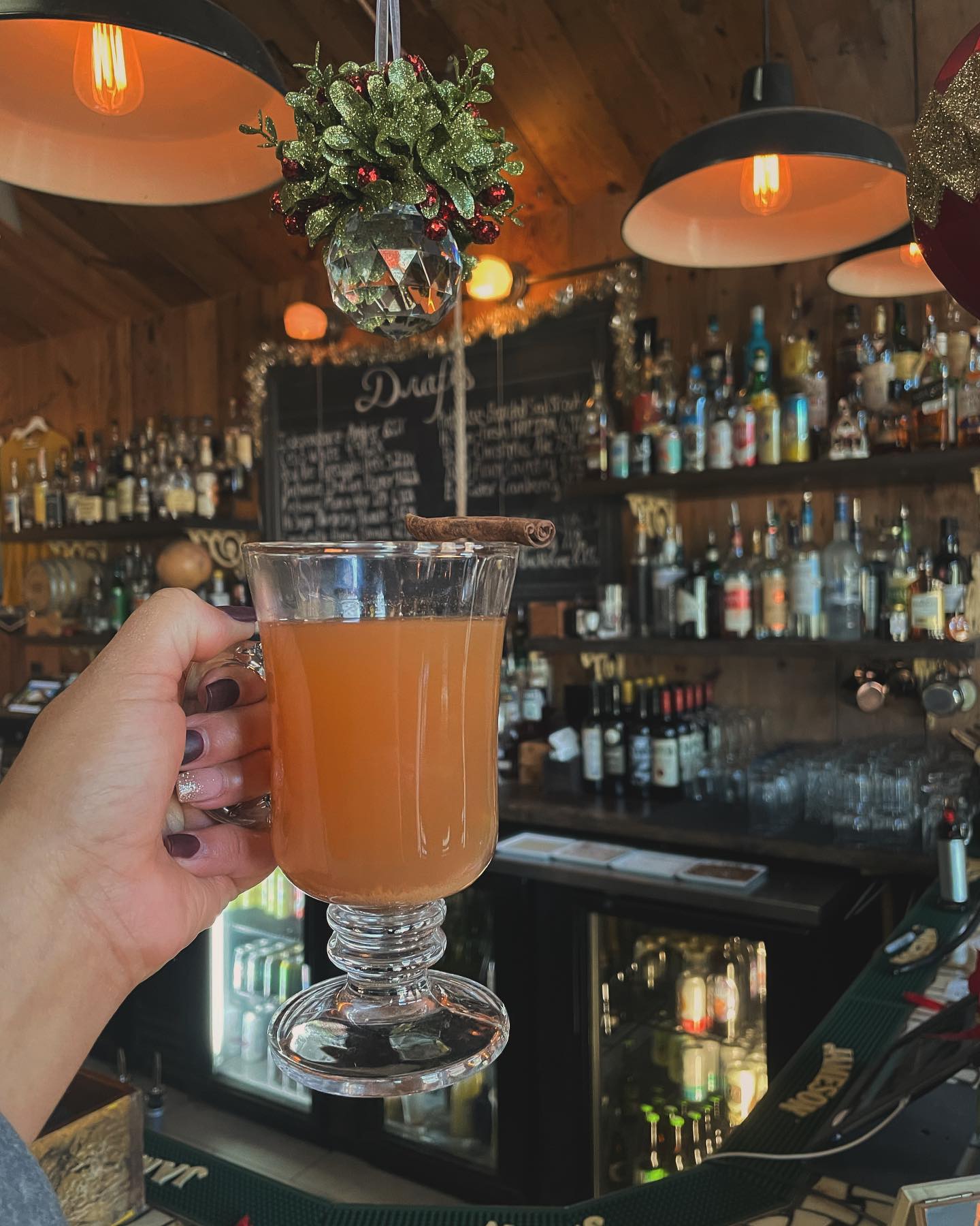 Hand holds an amber drink with cinnamon stick at a bar.