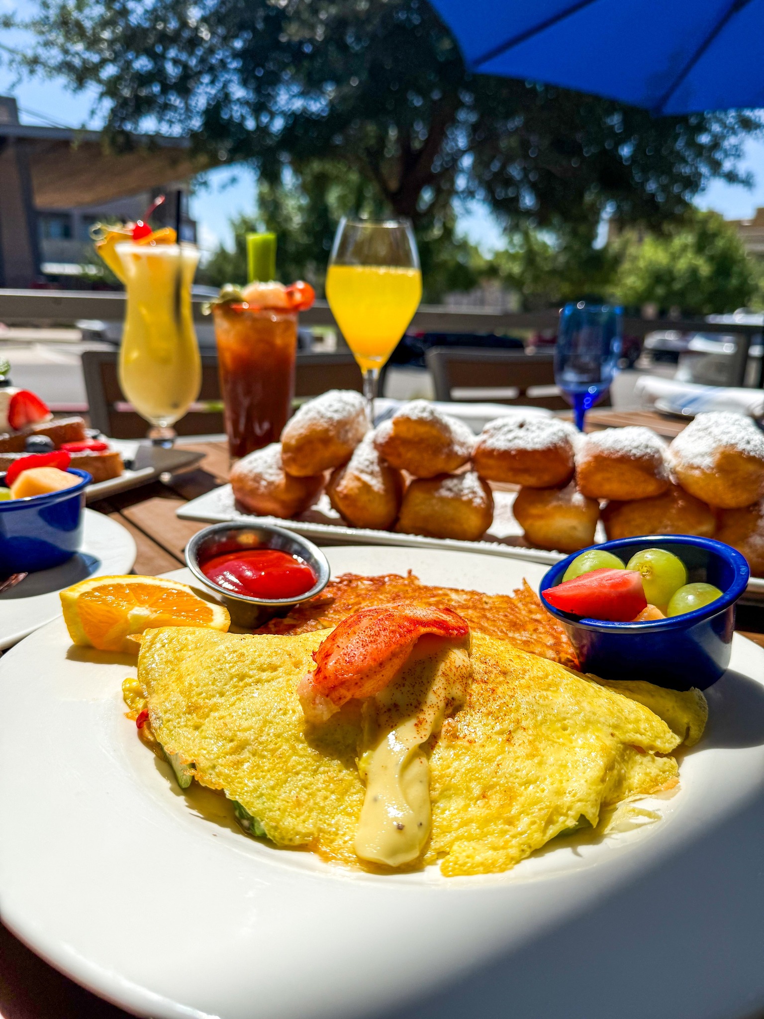 Outdoor brunch with lobster omelet, beignets, and various drinks.