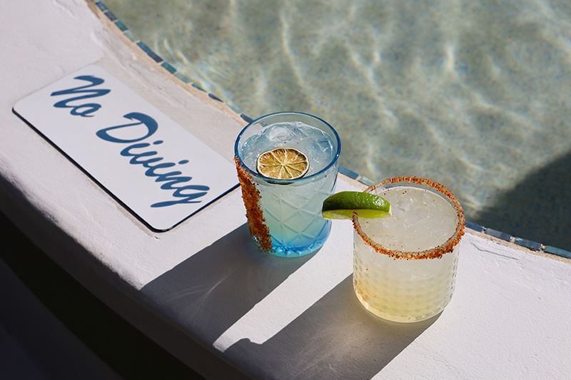 Two lime-garnished cocktails by a pool with a 'No Diving' sign.