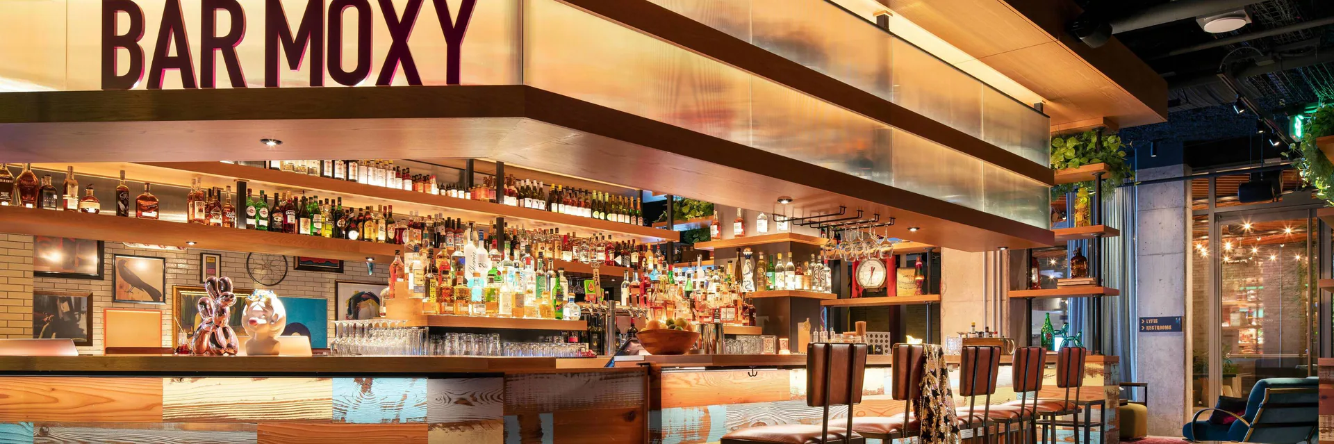 A well-lit, well-stocked BAR MOXY with numerous bottles and stools.
