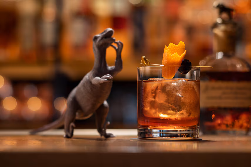 Toy dinosaur next to a cocktail with orange peel and cherry.
