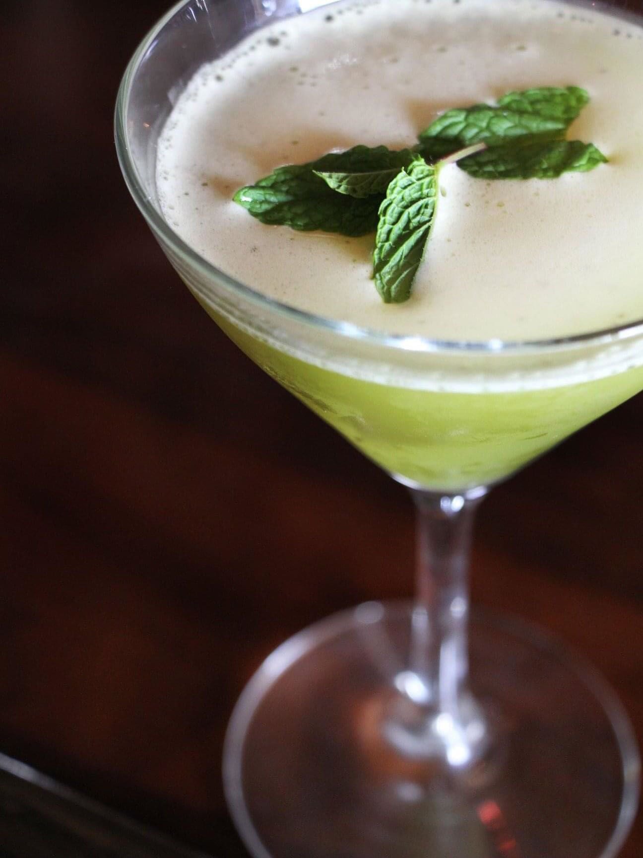 Green cocktail in martini glass with white foam and mint.