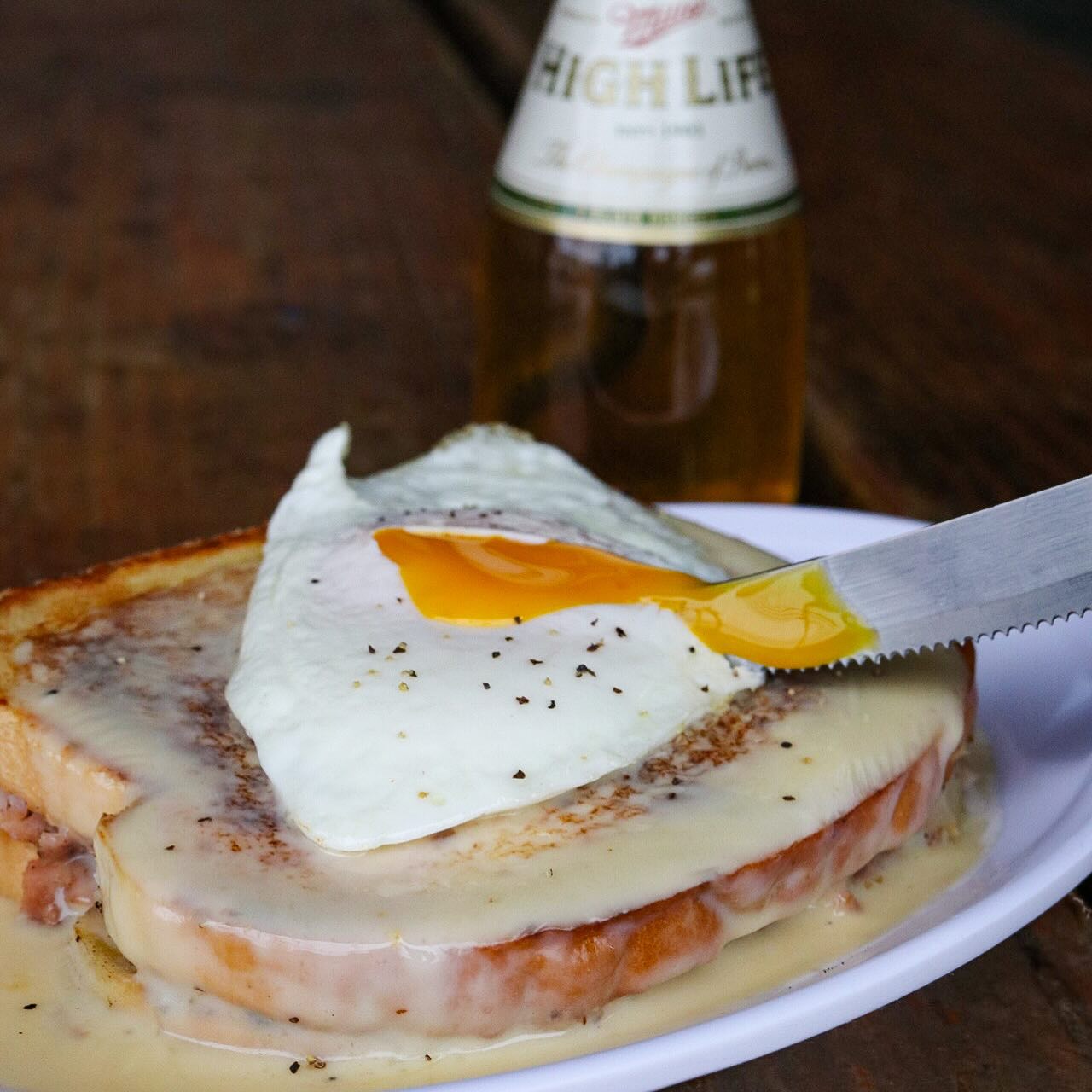 Knife cuts into runny fried egg on sauce-covered sandwich. Beer nearby.