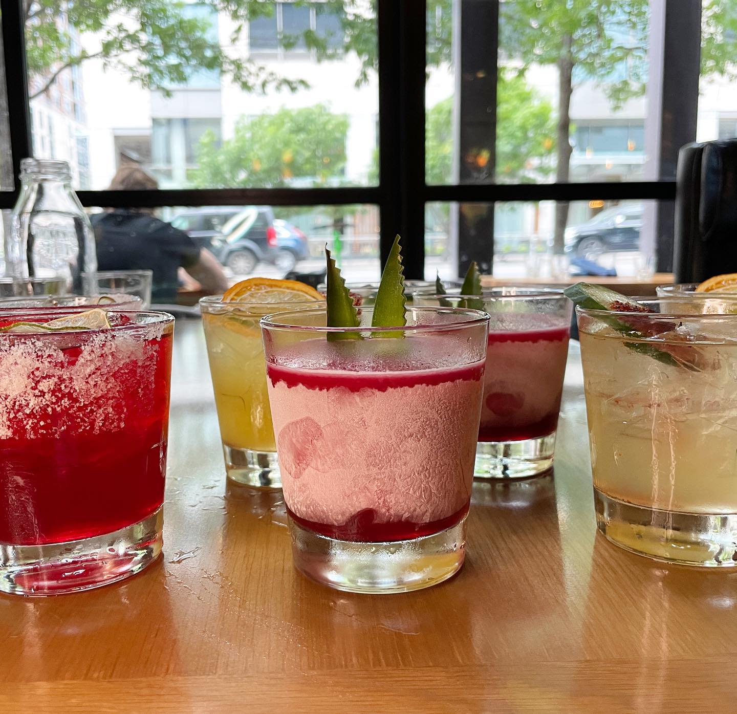 Multiple colorful drinks on a wooden table near a window.