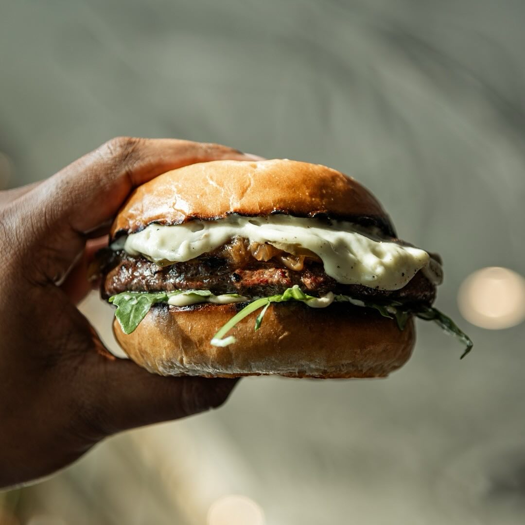 Hand holds a burger with melted cheese, caramelized onions, and arugula.