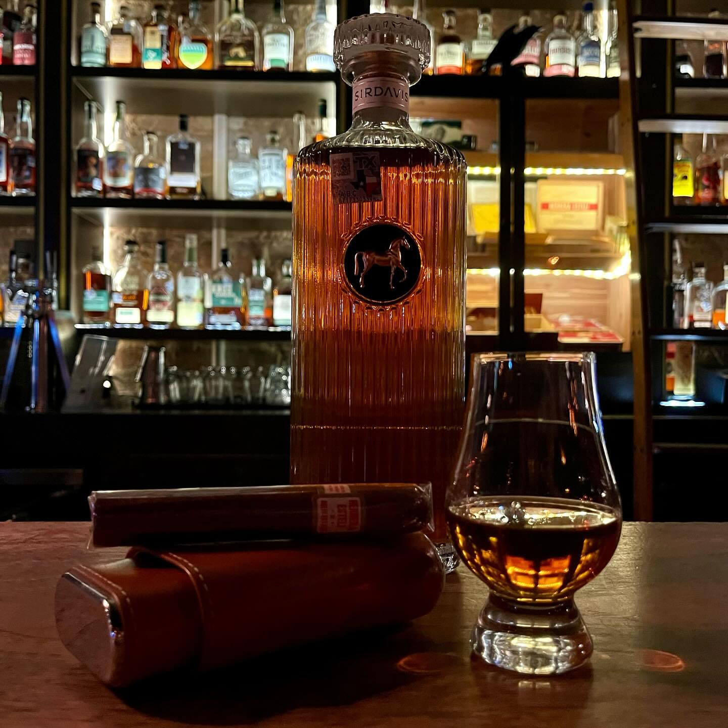 Tall liquor bottle, glencairn glass, cigar, and case on bar.