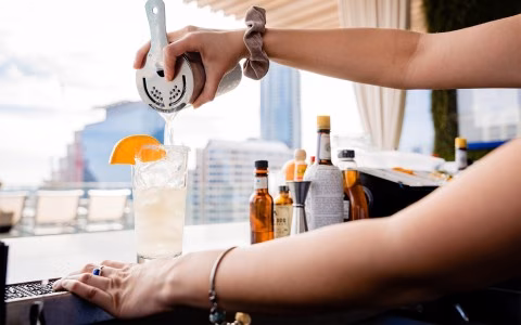 Person pouring drink from shaker into glass with orange slice.