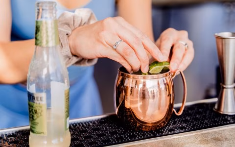 Hands adding lime wedges to a copper mug, ginger beer bottle present.