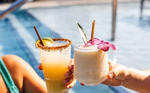 Hands holding two garnished cocktails by a bright blue swimming pool.