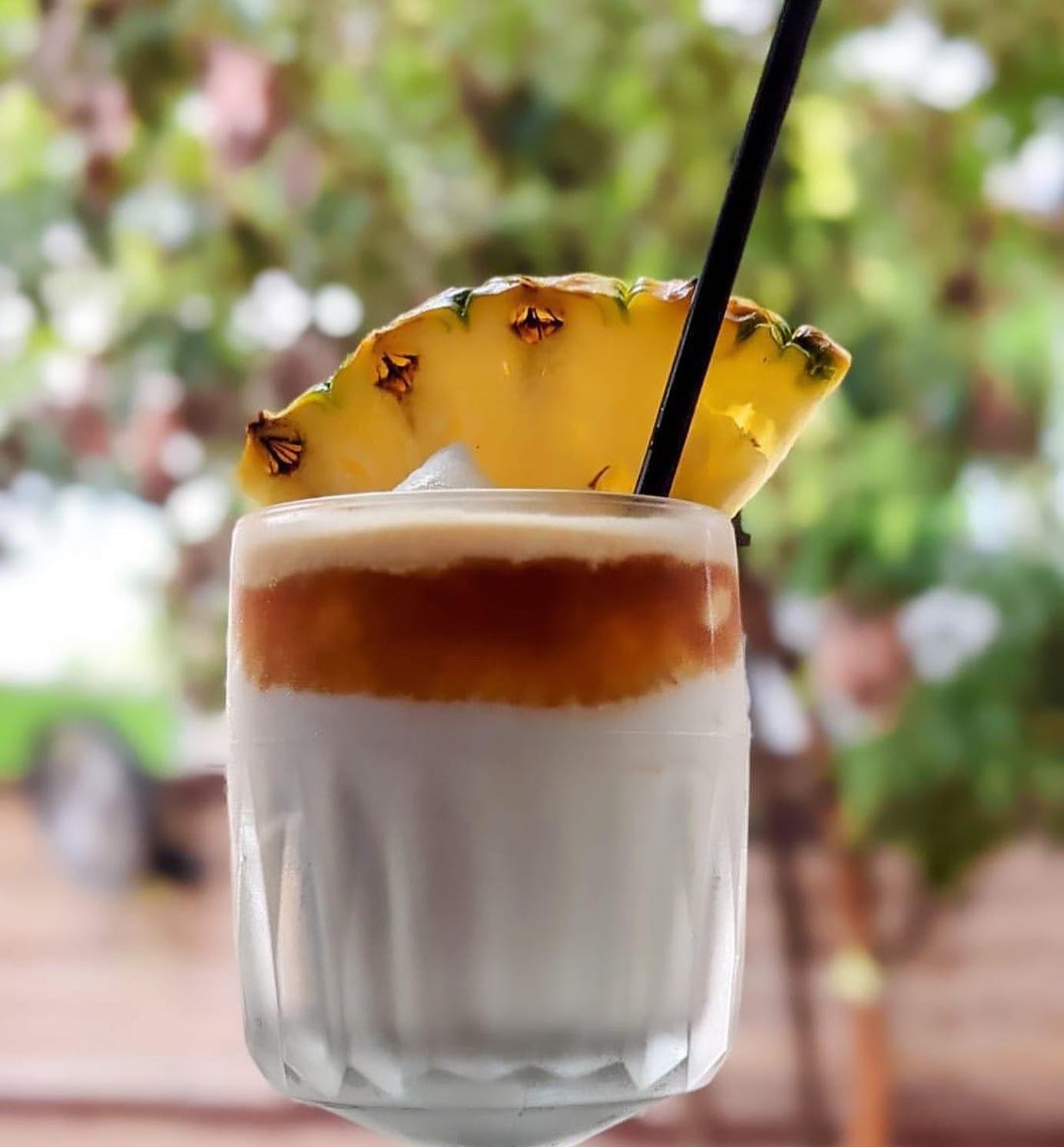 Layered drink with pineapple slice and straw, blurred green background.
