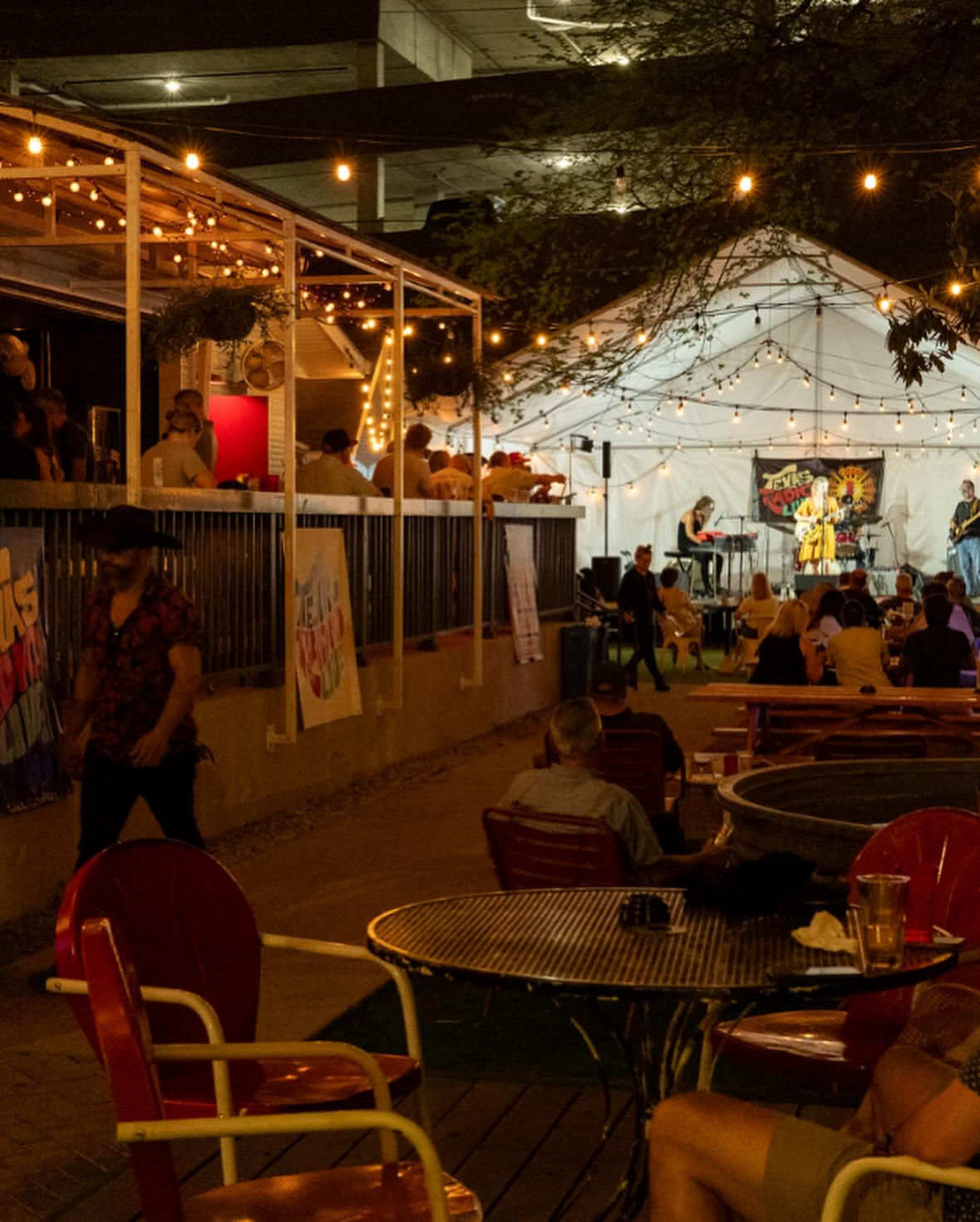 Night scene: outdoor bar and tent with live band and string lights.