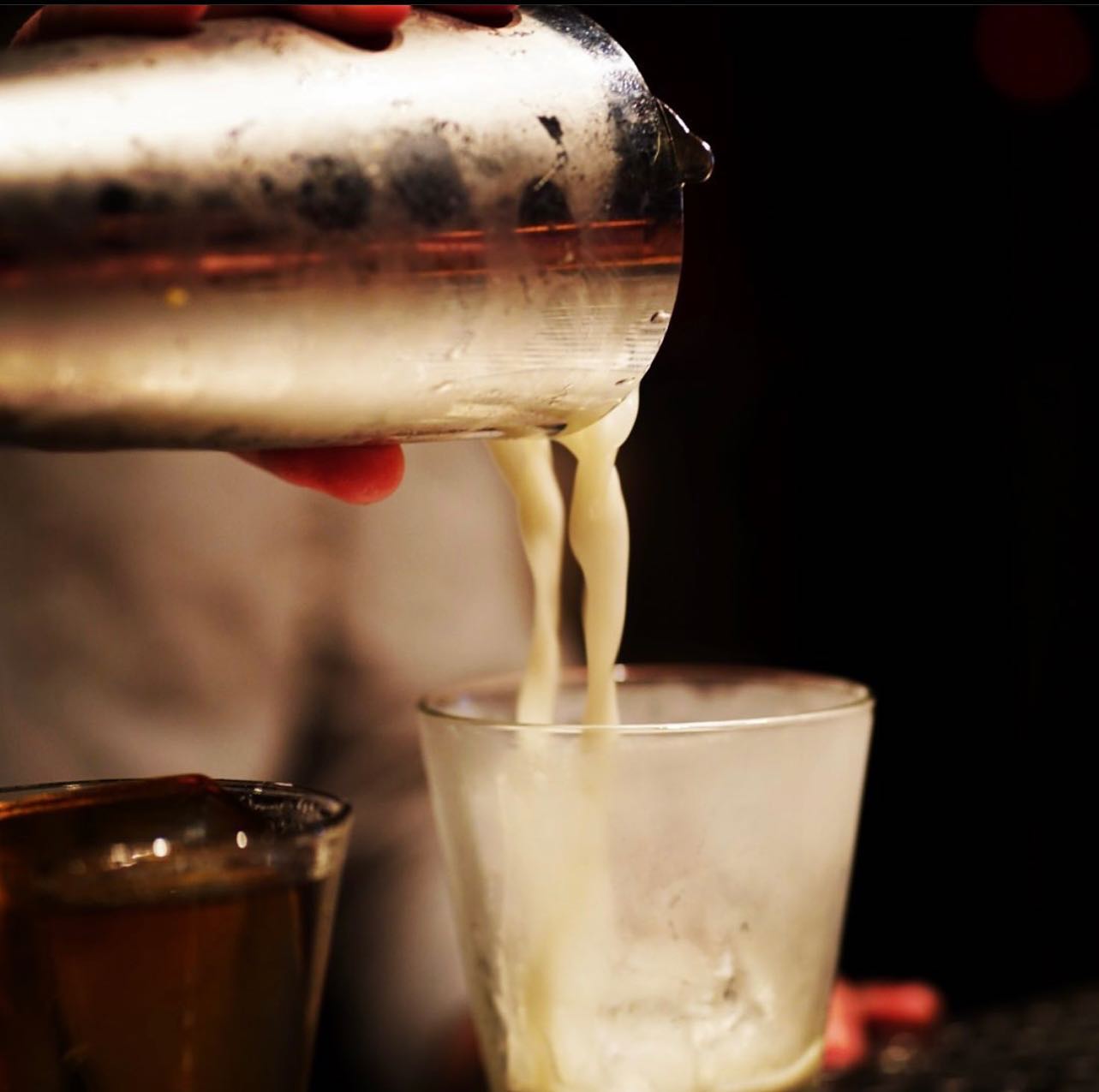 Hand pours light liquid from cocktail shaker into clear glass.
