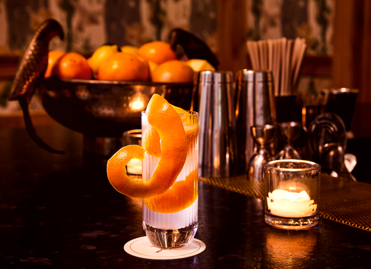 Cocktail with swirled orange peel, ice, and candle on dark bar.