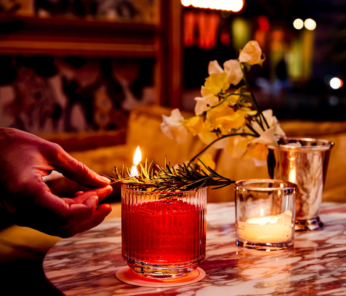 Hand flaming rosemary over a red cocktail on a marble table.