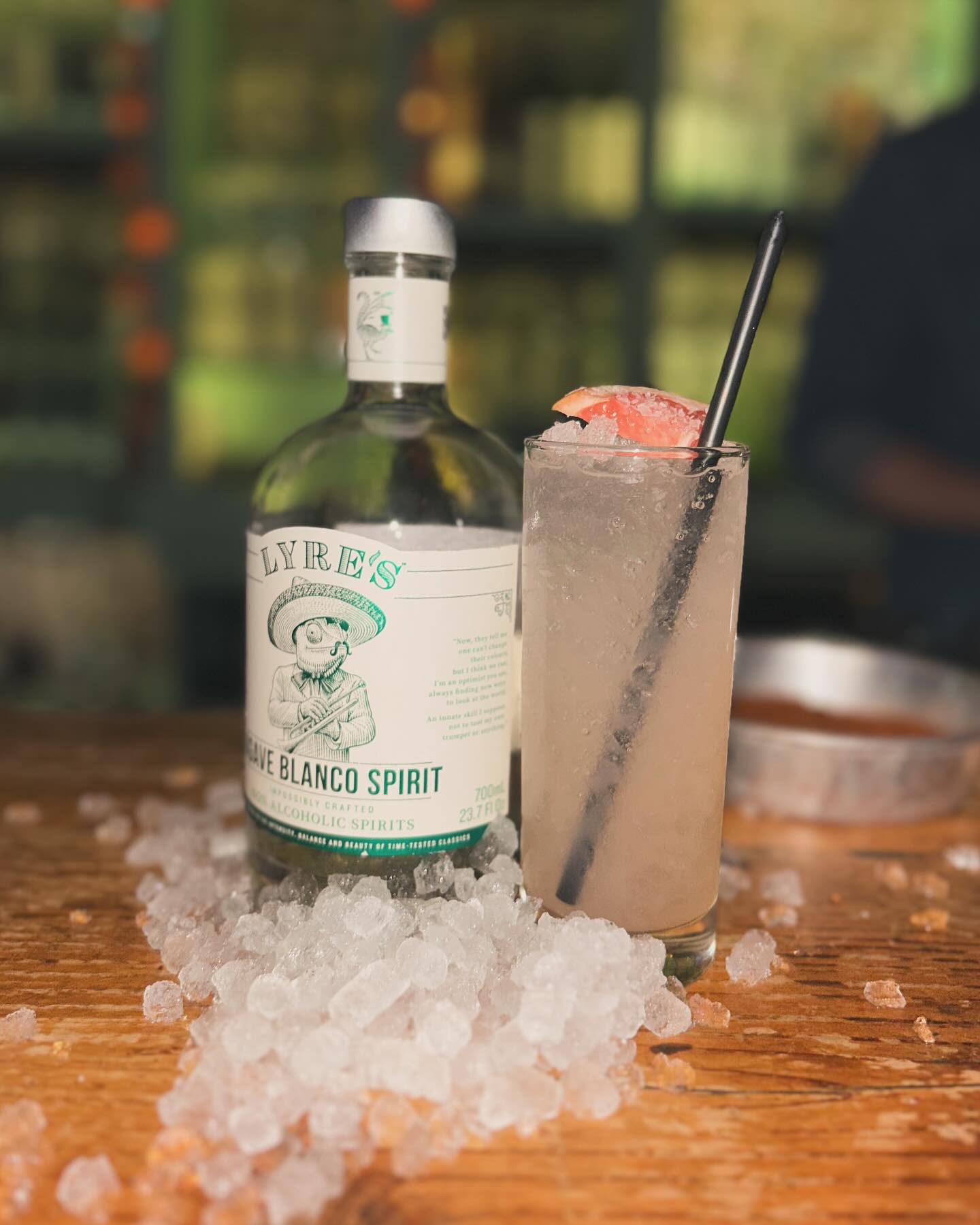 Lyre's Agave Blanco bottle, a grapefruit cocktail, and crushed ice on wood.