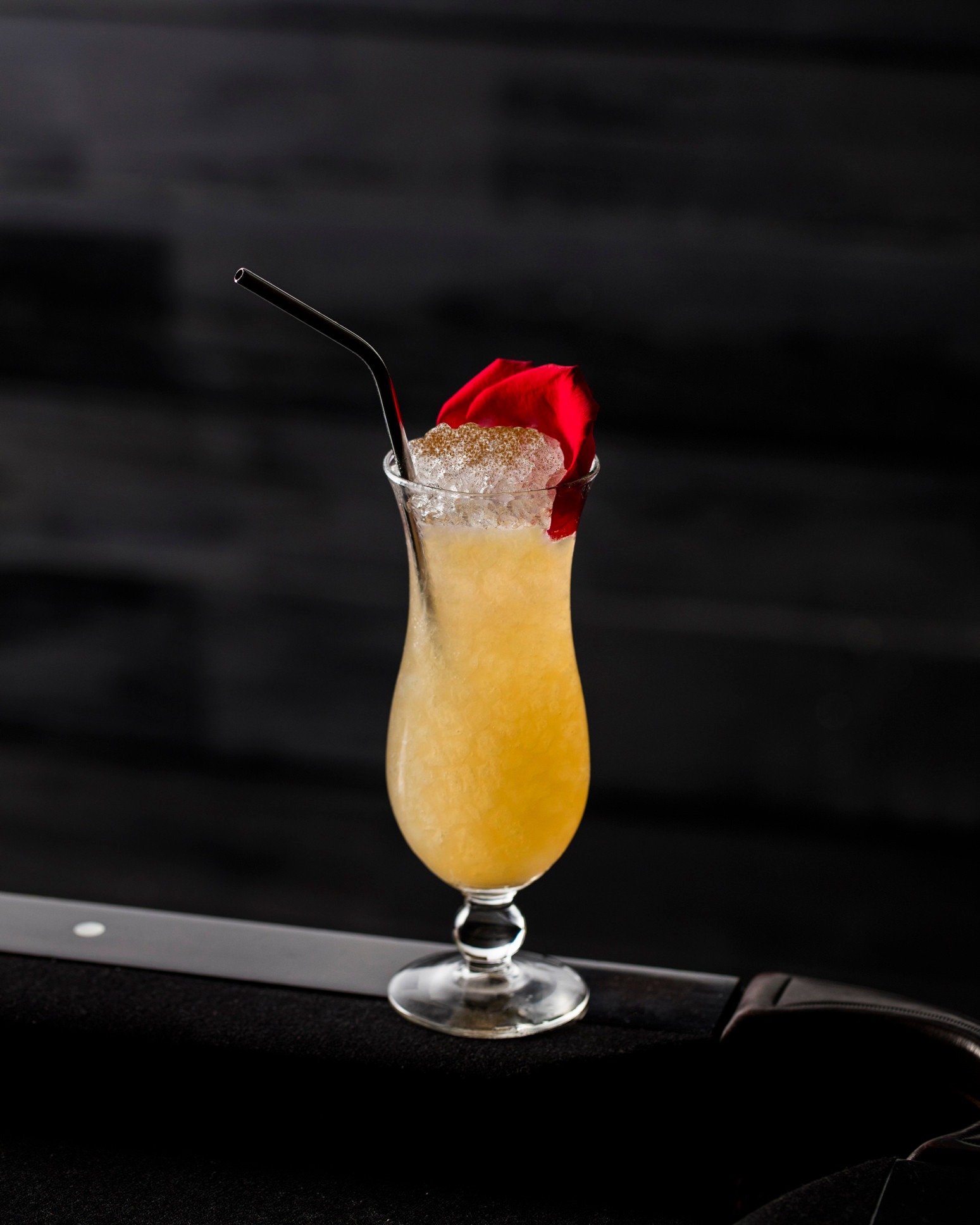 Yellow cocktail with crushed ice, red rose petal, and dark straw.