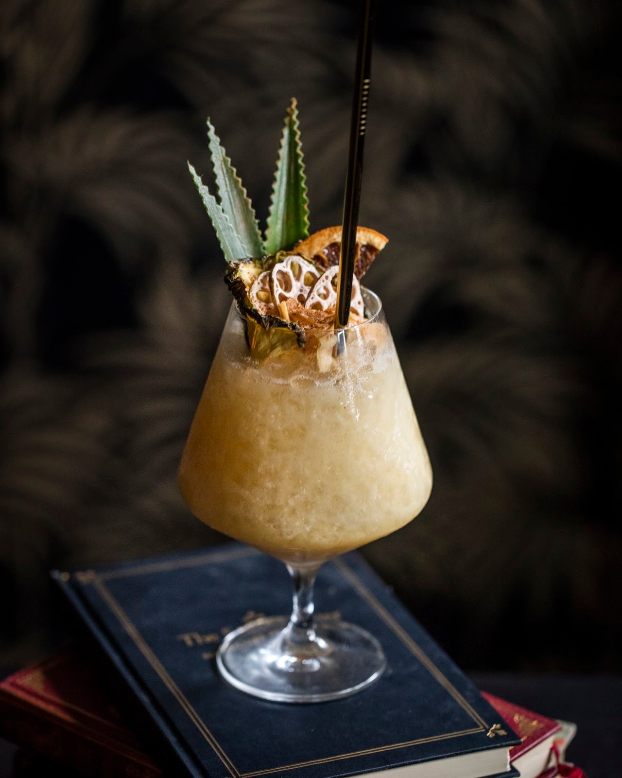 Frosted cocktail garnished with pineapple, dried fruit, and a straw on books.