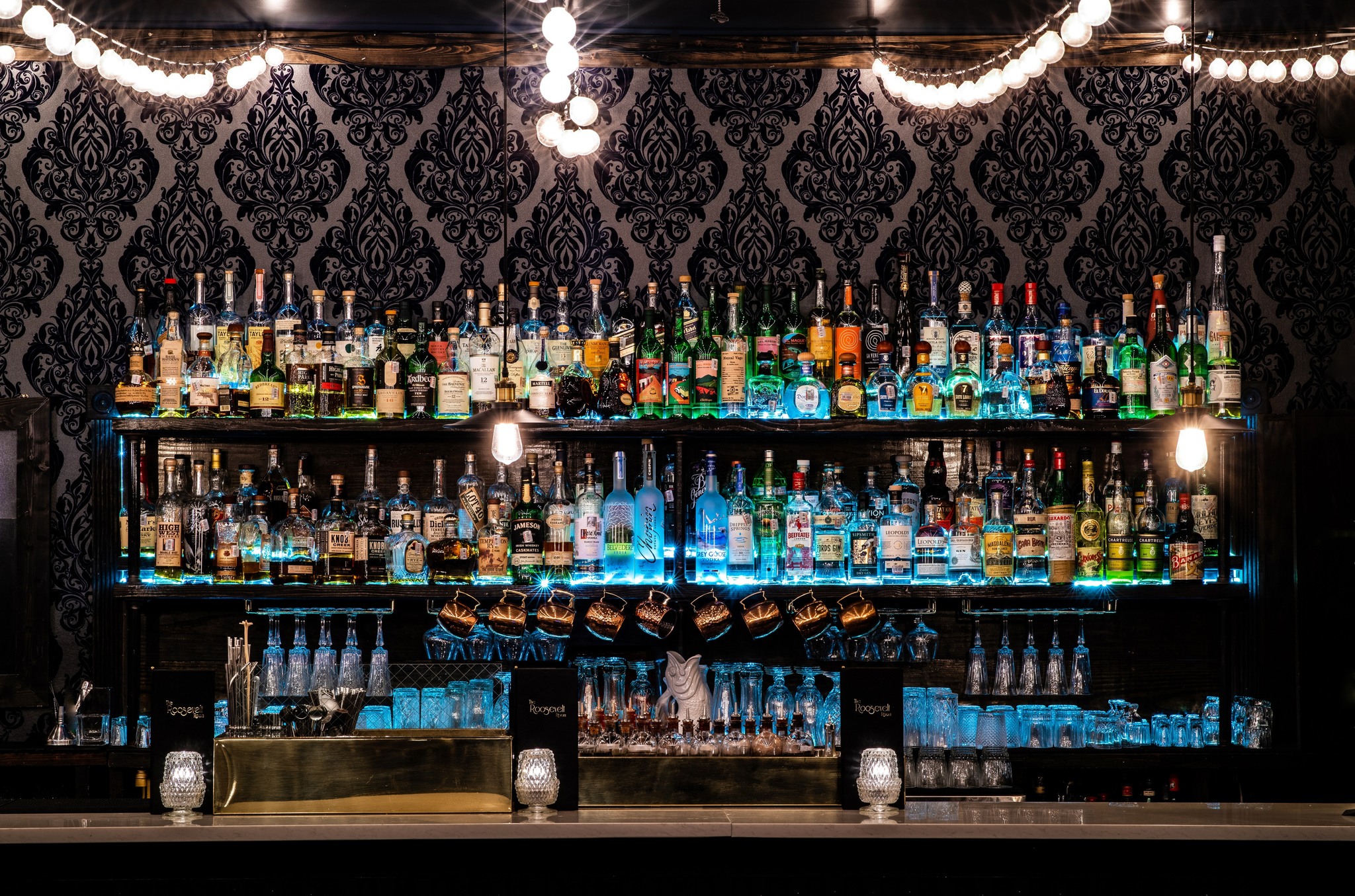 Many liquor bottles on illuminated bar shelves with patterned wallpaper.