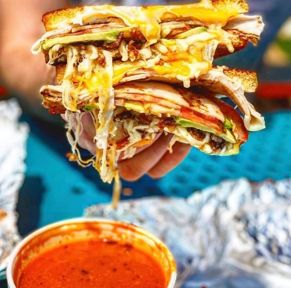 Large sandwich with meat, avocado, dripping cheese held above red liquid.