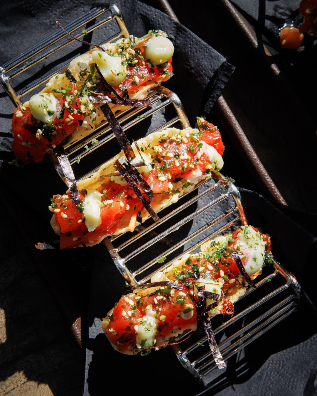 Three appetizers with red cubes, rice, herbs, and seaweed on racks.