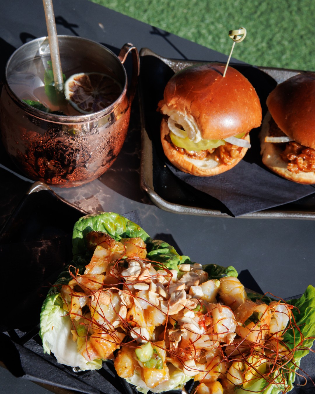 Copper mug, two sliders, and shrimp lettuce wraps on a dark table.