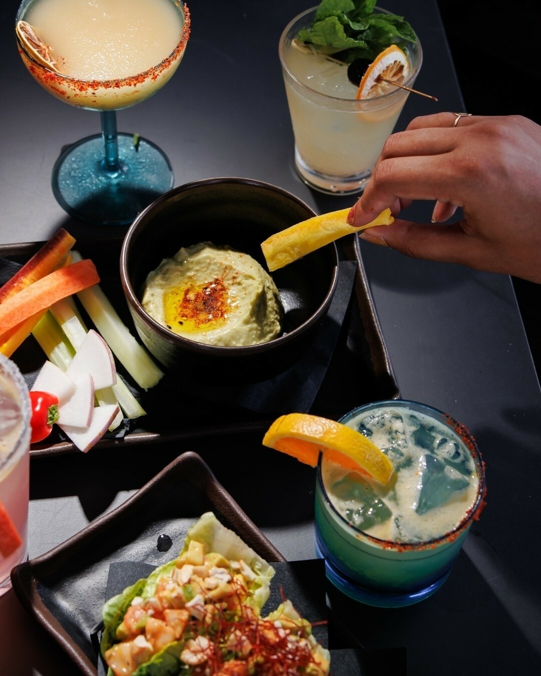 Hand dipping a fry into green dip, with cocktails and snacks.