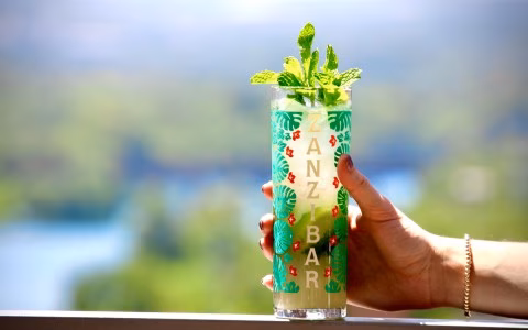 Hand holding a mint drink in a Zanzibar-branded glass outdoors.