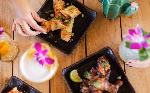A hand picks an egg roll from a table with wings and drinks.