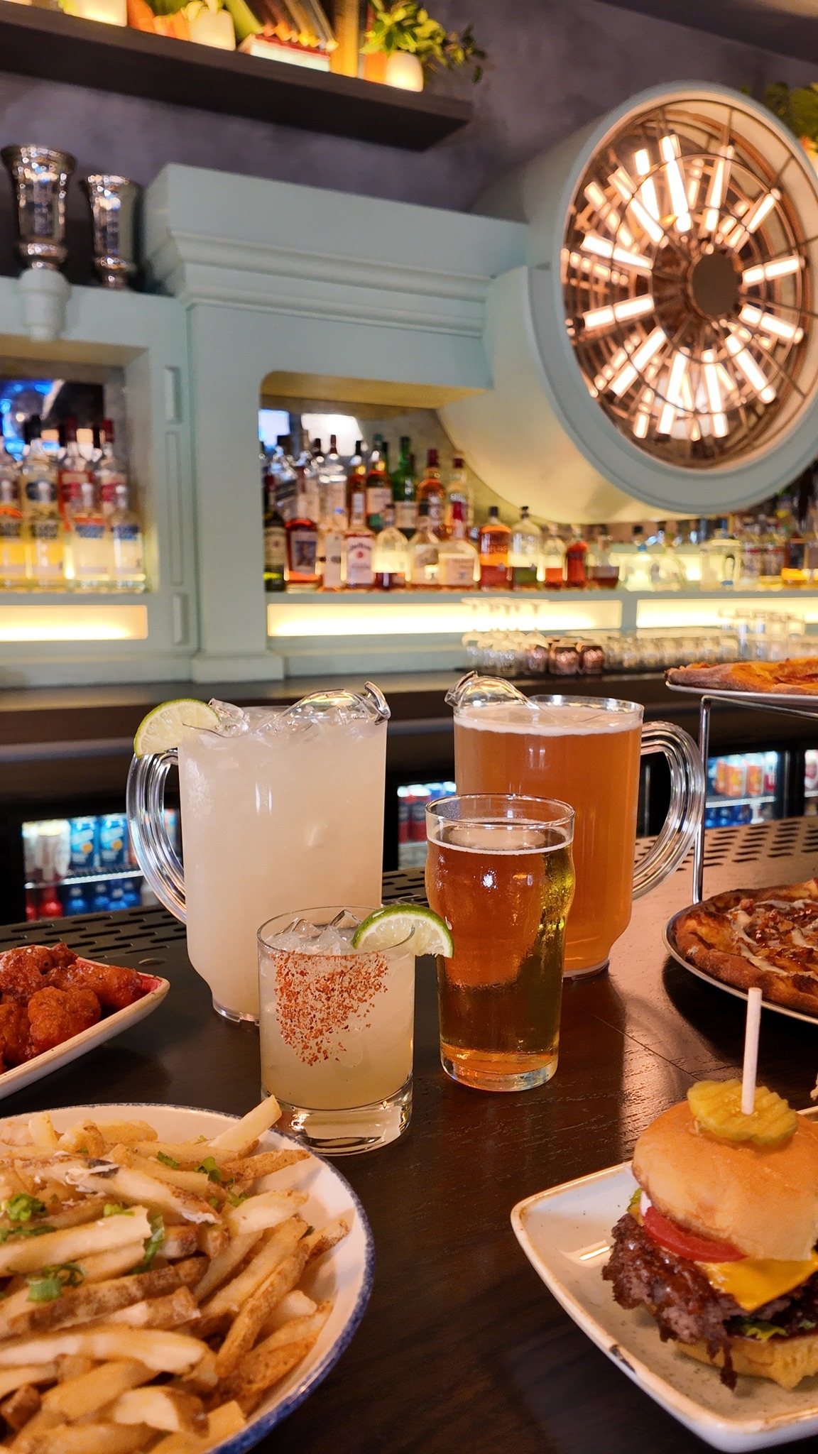Bar table with pitchers, glasses, fries, burger, wings, and pizza.