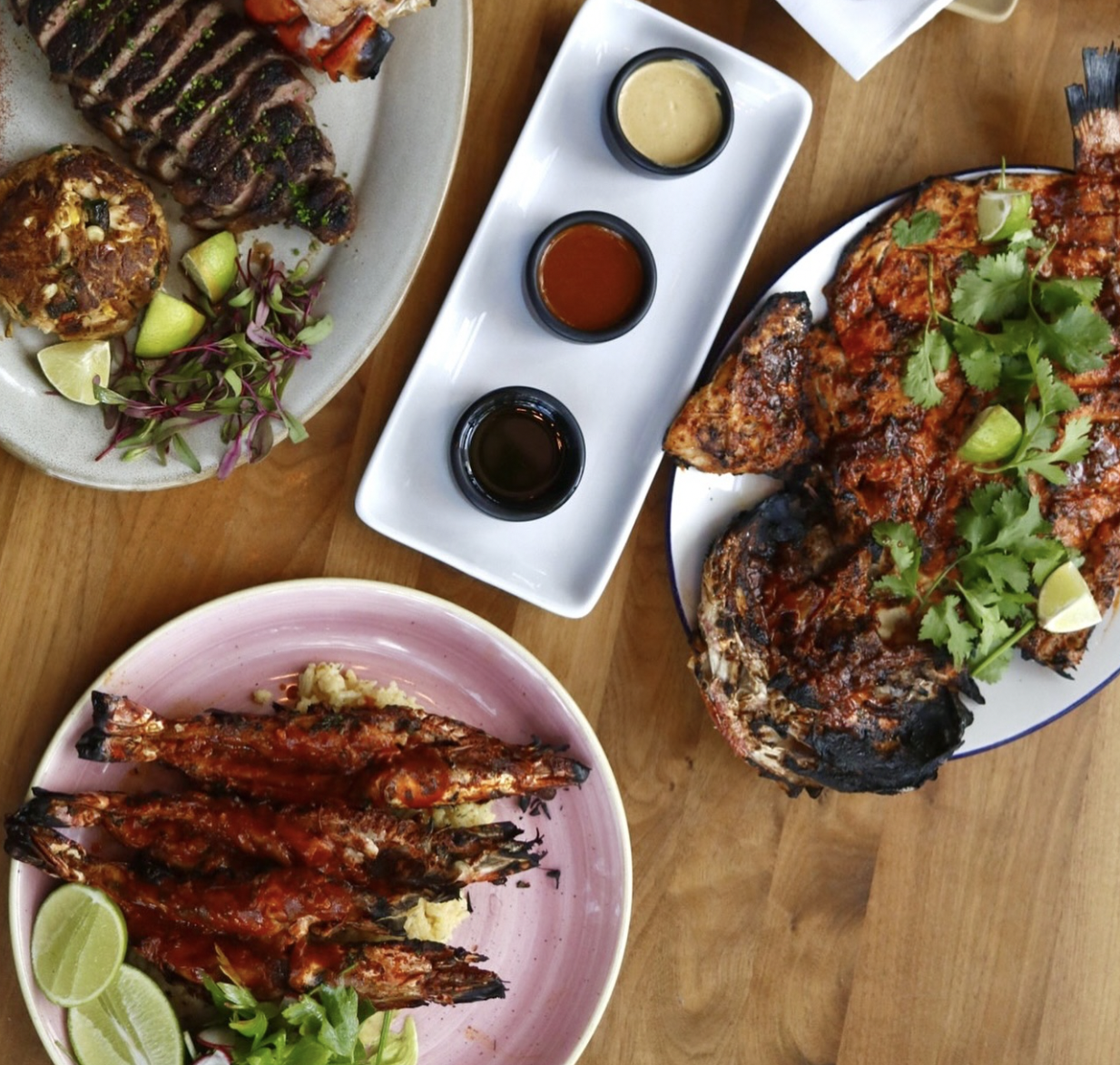 Overhead view of grilled fish, shrimp, steak, and sauces on a table.