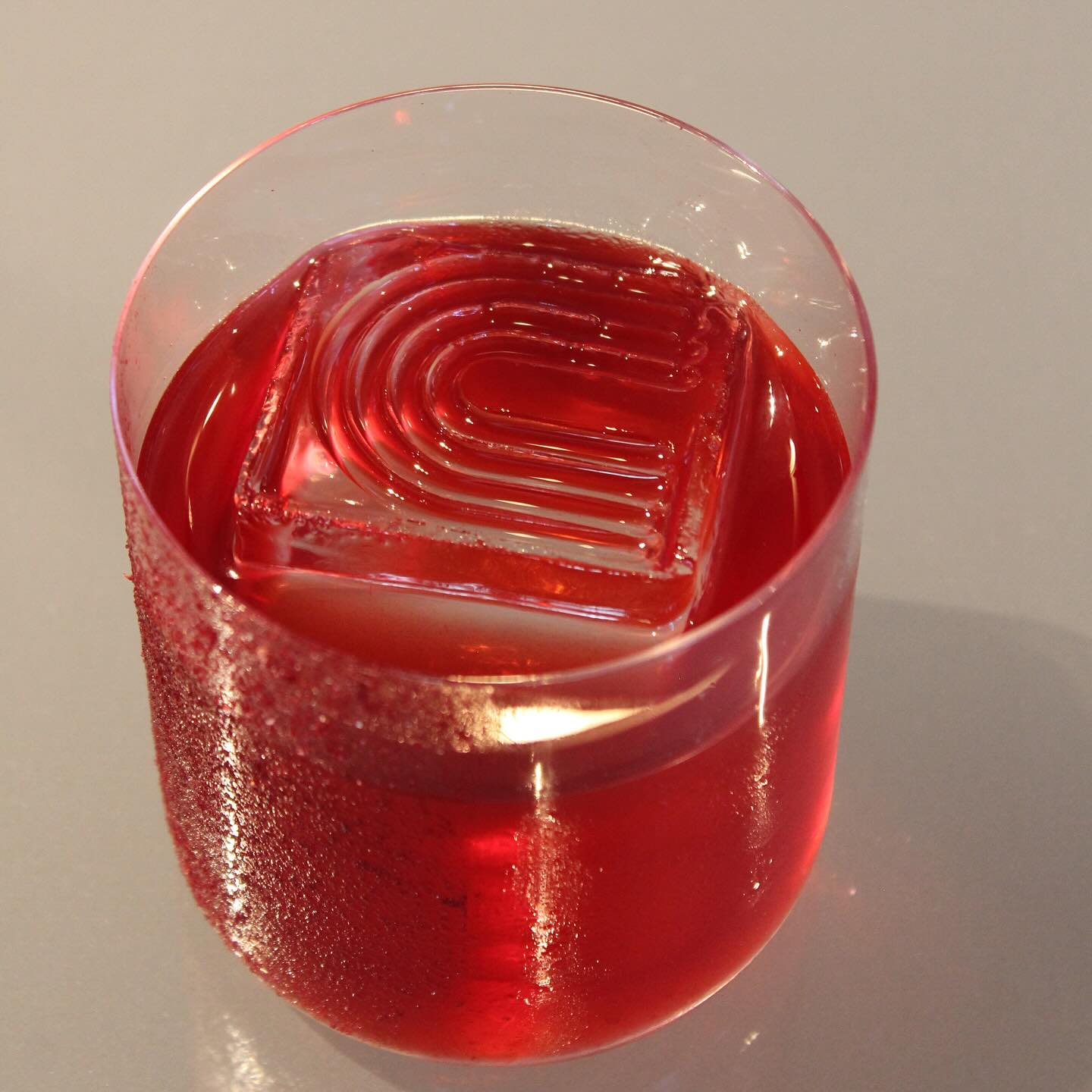 Red liquid in glass with square ice cube featuring a molded pattern.