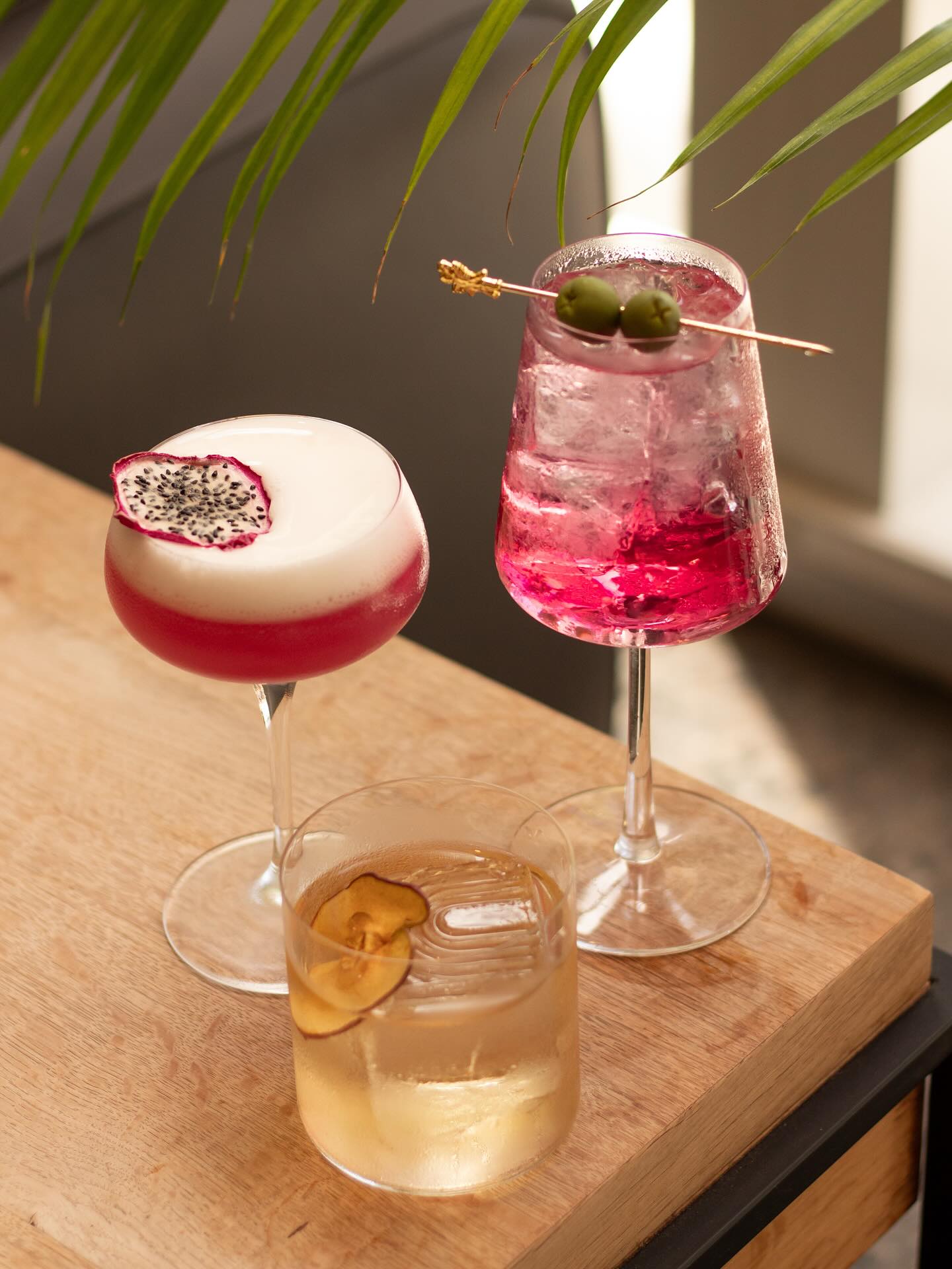 Three colorful cocktails on a wooden table with palm leaves.